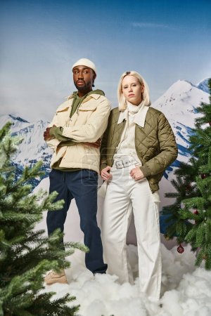 fashionable couple in warm attire posing next to fir trees with mountain backdrop, diverse couple