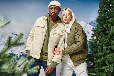 stylisches multiethnisches Paar in warmer Winterkleidung posiert gemeinsam vor verschneiter Kulisse, Modekonzept