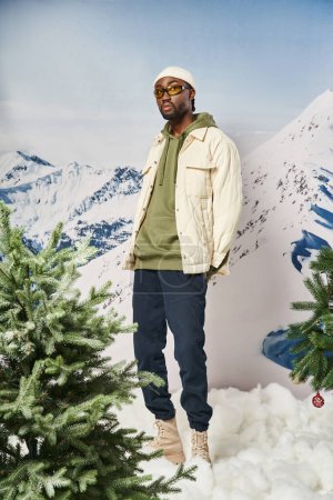 fashionable man in stylish warm outfit with beanie hat posing next to pine trees, winter concept
