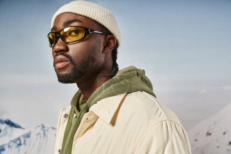 hombre afroamericano guapo en gorro blanco sombrero y gafas de sol mirando a la cámara, moda de invierno