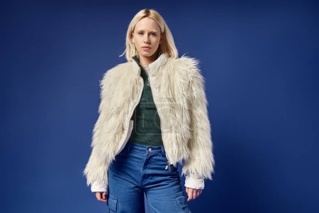 stylish blonde woman in white faux fur jacket and denim jeans looking at camera on blue background