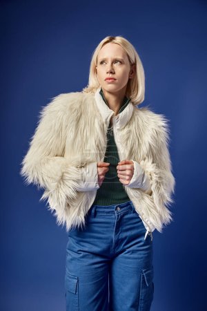 pensive and stylish blonde woman in faux fur jacket and denim jeans posing on blue backdrop