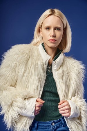 Wintermode, stylische blonde Frau in Kunstpelzjacke und Jeans posiert vor blauem Hintergrund