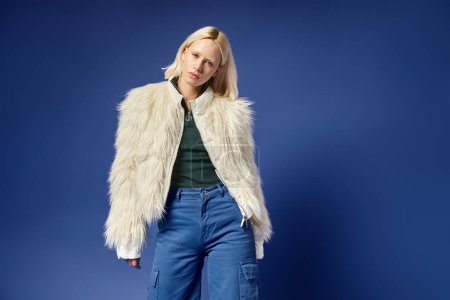 winter style, attractive young woman in faux fur jacket and denim jeans posing on blue backdrop
