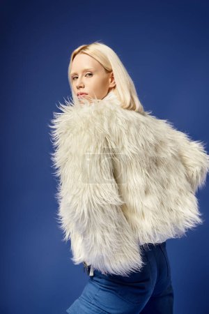 winter attire, attractive young woman in faux fur jacket and denim jeans posing on blue backdrop