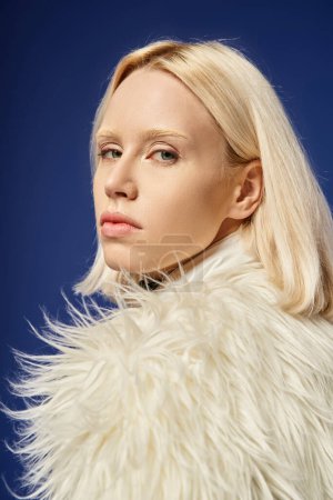 winter style, portrait of young woman in white faux fur jacket looking at camera on blue backdrop