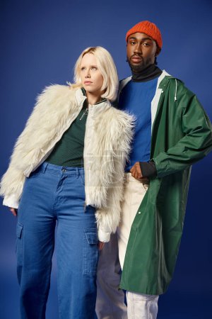 stilvolles gemischtrassiges Paar in Winterkleidung vor blauem Hintergrund, blonde Frau und afrikanisch-amerikanischer Mann