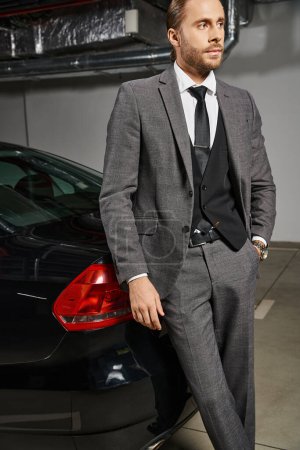 attraktiver Geschäftsmann mit Bart im schicken Anzug und posiert neben seinem Auto auf einem Parkplatz