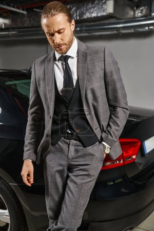 gut aussehender raffinierter Mann im eleganten Anzug, der in der Nähe seines Autos mit der Hand in der Tasche posiert, Geschäftskonzept