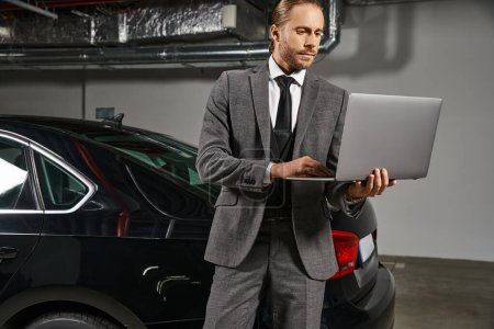 gut aussehender Mann im schicken Anzug arbeitet am Laptop auf dem Parkplatz in der Nähe seines Autos, Geschäftskonzept