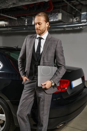 schöner Profi im schicken Anzug mit Krawatte, Laptop in der Hand und posiert vor seinem Auto, Geschäft