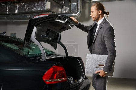 attraktiver stylischer Geschäftsmann im eleganten schicken Anzug mit Pferdeschwanz im Kofferraum seines Autos