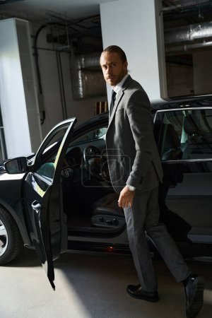 Foto de Apuesto hombre de negocios con barba y cola de caballo de pie junto a su coche y mirando a la cámara - Imagen libre de derechos
