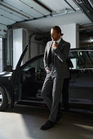 Foto de Atractivo hombre barbudo en traje elegante posando junto a su coche con la mano en su corbata, concepto de negocio - Imagen libre de derechos