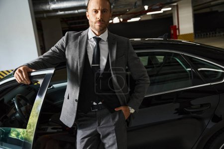 Foto de Hombre de negocios guapo con barba posando cerca de su coche con la mano en el bolsillo y mirando a la cámara - Imagen libre de derechos