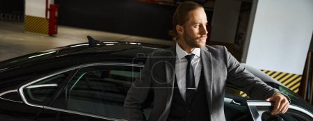 Foto de Atractivo hombre barbudo en traje inteligente posando cerca de su coche y mirando hacia otro lado, concepto de negocio, pancarta - Imagen libre de derechos
