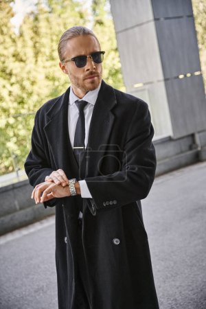 attraktiver eleganter Geschäftsmann in schwarzem Mantel mit Sonnenbrille, der draußen mit seiner Armbanduhr posiert