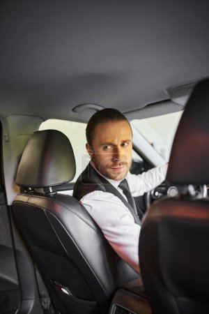 Foto de Atractivo hombre barbudo en chaleco negro mirando hacia atrás mientras está detrás del volante, concepto de negocio - Imagen libre de derechos