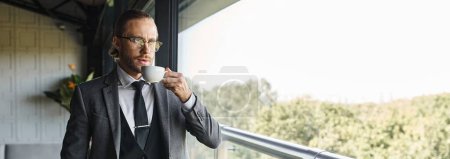 hombre de negocios bien parecido en traje inteligente con gafas de beber té y mirando por la ventana, pancarta