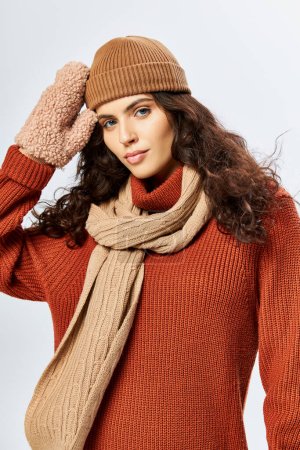 curly woman in hat and knitted terracotta sweater posing in scarf and mittens on grey backdrop