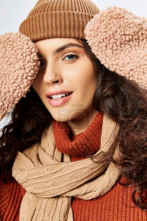 cheerful woman in hat and knitted terracotta sweater posing in scarf and mittens on grey backdrop