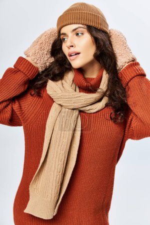 charming woman in hat and knitted terracotta sweater posing in scarf and mittens on grey backdrop