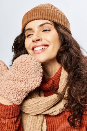 smiling woman in hat and knitted terracotta sweater posing in scarf and mittens on grey backdrop
