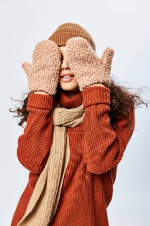 young woman in hat and knitted terracotta sweater covering eyes with mittens on grey backdrop