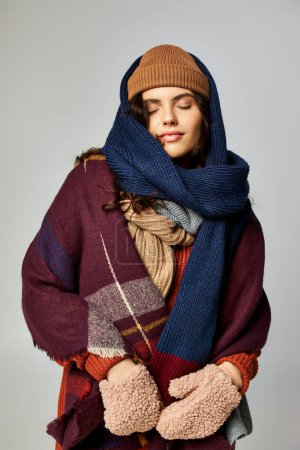 estilo invierno, mujer joven en capas de ropa, sombrero de punto y bufandas posando sobre fondo gris