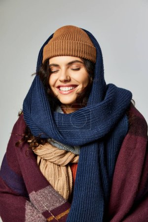 winter fashion, cheerful woman in layered clothing, knitted hat and scarfs posing on grey backdrop