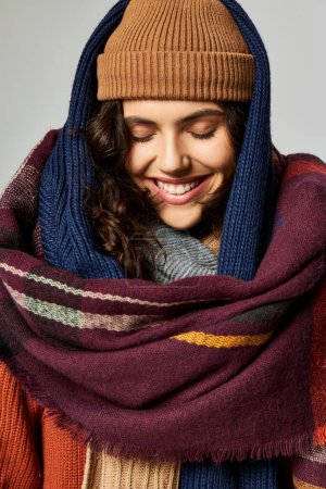 moda de invierno, mujer alegre en capas de ropa, sombrero de punto y bufandas posando sobre fondo gris