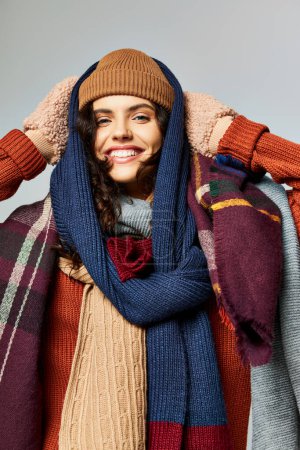 moda de invierno, mujer positiva en capas de ropa, sombrero de punto y bufandas posando sobre fondo gris