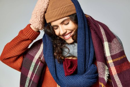 moda de invierno, mujer alegre en capas de ropa, sombrero caliente y bufandas posando sobre fondo gris