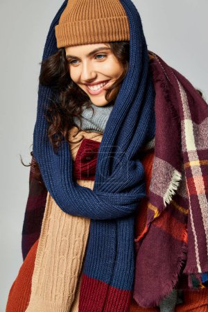 estilo invierno, mujer morena feliz en capas de ropa, sombrero caliente y bufandas posando sobre fondo gris