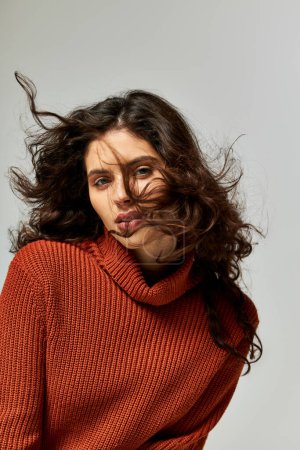 wind blowing at face of brunette curly woman in terracotta sweater on grey backdrop, winter fashion