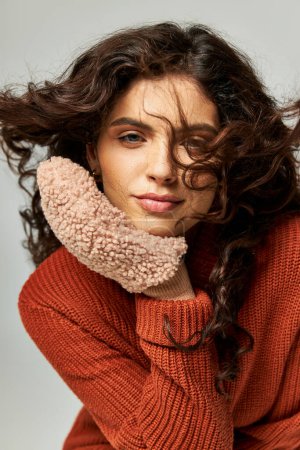 wind blowing at face of curly brunette woman in terracotta sweater and mittens, winter fashion