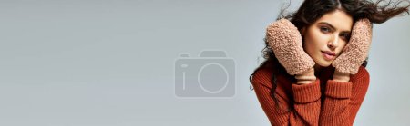 curly brunette woman in terracotta sweater and soft mittens posing on grey, winter fashion banner