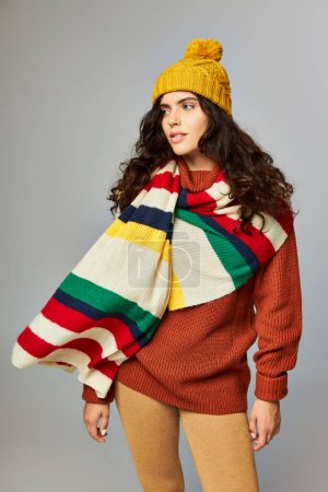 curly woman in knitted bobble hat and sweater with stripped scarf on top posing on grey backdrop