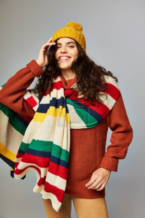 happy young woman in knitted bobble hat and sweater with stripped scarf posing on grey background