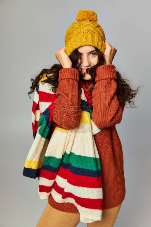 happy woman wearing cozy bobble hat and sweater with stripped scarf posing on grey background