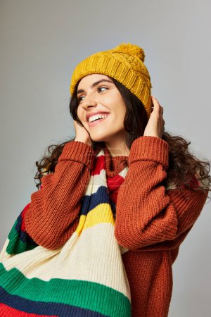 pleased woman wearing cozy bobble hat and sweater with stripped scarf posing on grey background