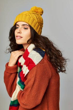 brunette woman with curly hair in bobble hat and sweater with stripped scarf posing on grey