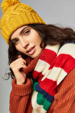 portrait of brunette woman with curly hair in bobble hat and sweater with stripped scarf on grey