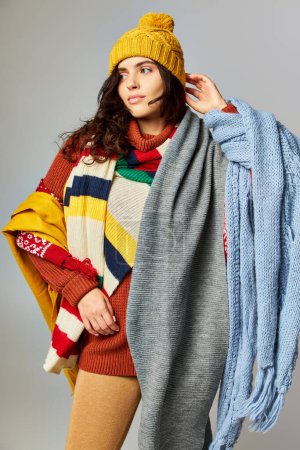 brunette woman with curly hair in bobble hat and cozy sweater posing with scarfs on grey backdrop