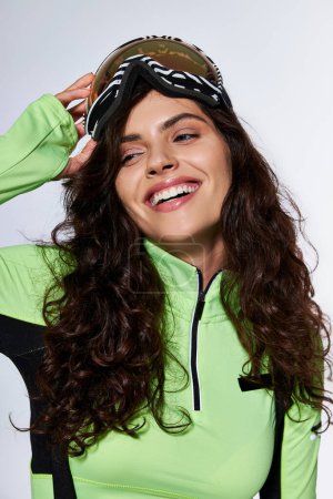 cheerful young woman with curly hair posing in stylish ski clothes and glasses on grey backdrop