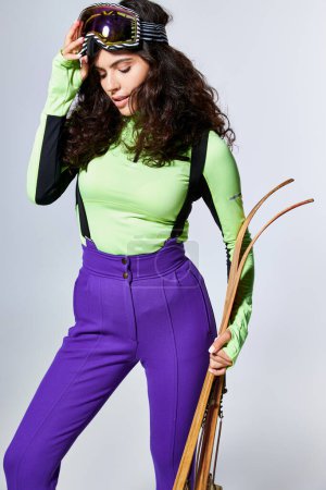 brunette young woman with curly hair posing in trendy sporty clothes and holding skis on grey