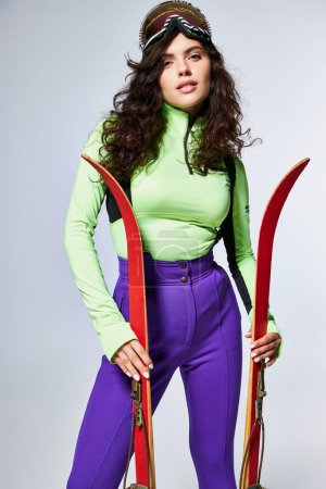 attraktive junge Frau mit lockigem Haar posiert in angesagter Active Wear und hält Skier auf grau