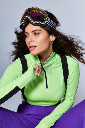 brunette sporty woman with curly hair zipping green long sleeve  while posing on grey backdrop