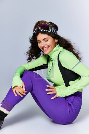 Wintersport, fröhliche und sportliche Frau mit lockigem Haar sitzt in aktiver Kleidung vor grauem Hintergrund
