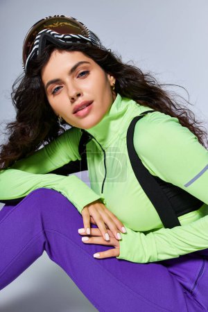 winter sport, beautiful and sporty woman with curly hair sitting in active wear on grey backdrop
